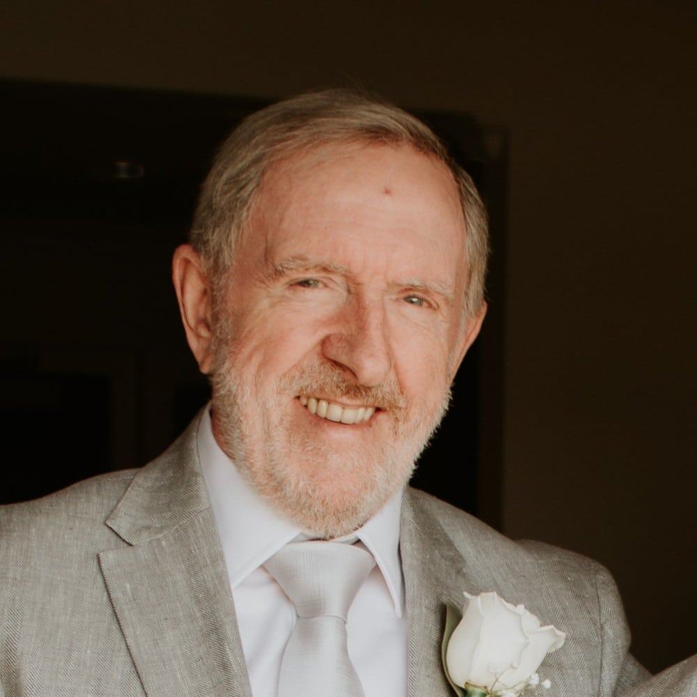 Friendly elderly man dressed in a gray suit with a white rose boutonniere, smiling warmly.