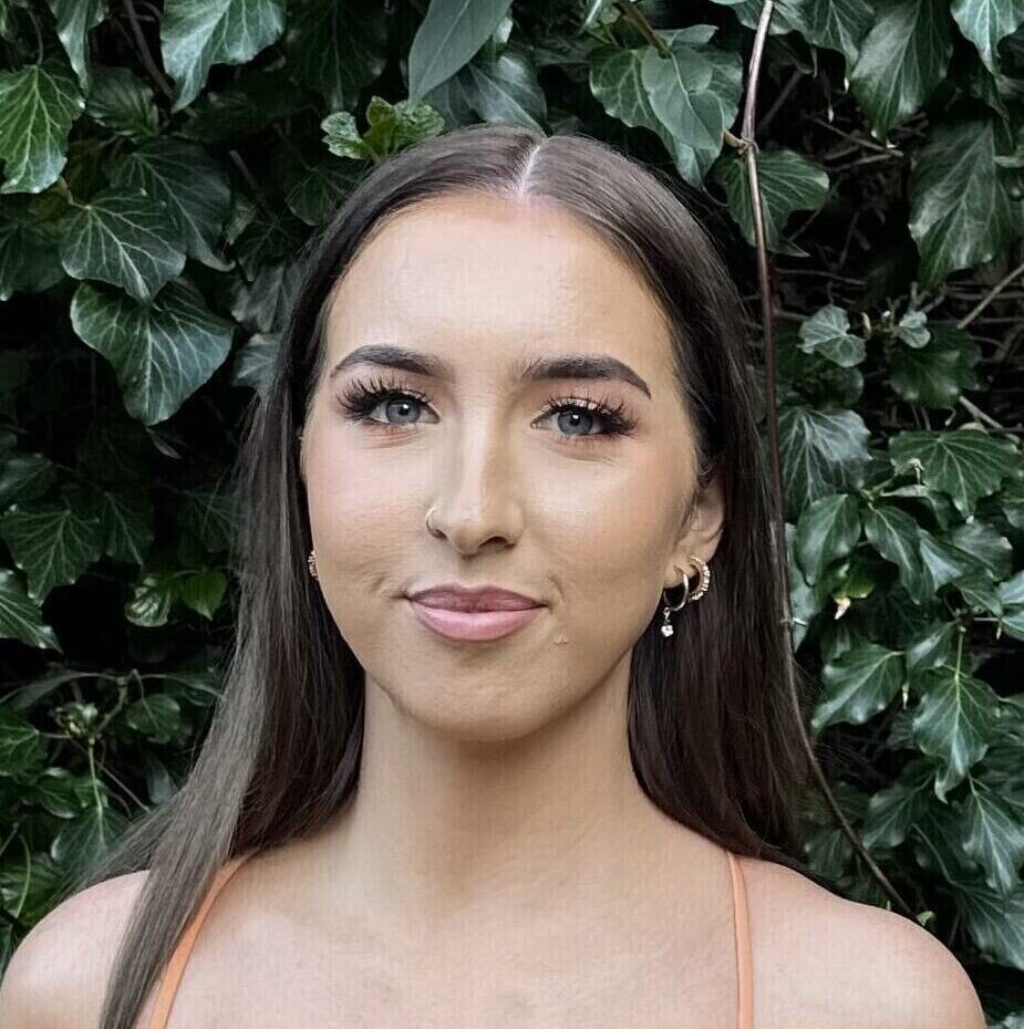 Portrait of a young woman with long dark hair and natural makeup, standing in front of dense green l.